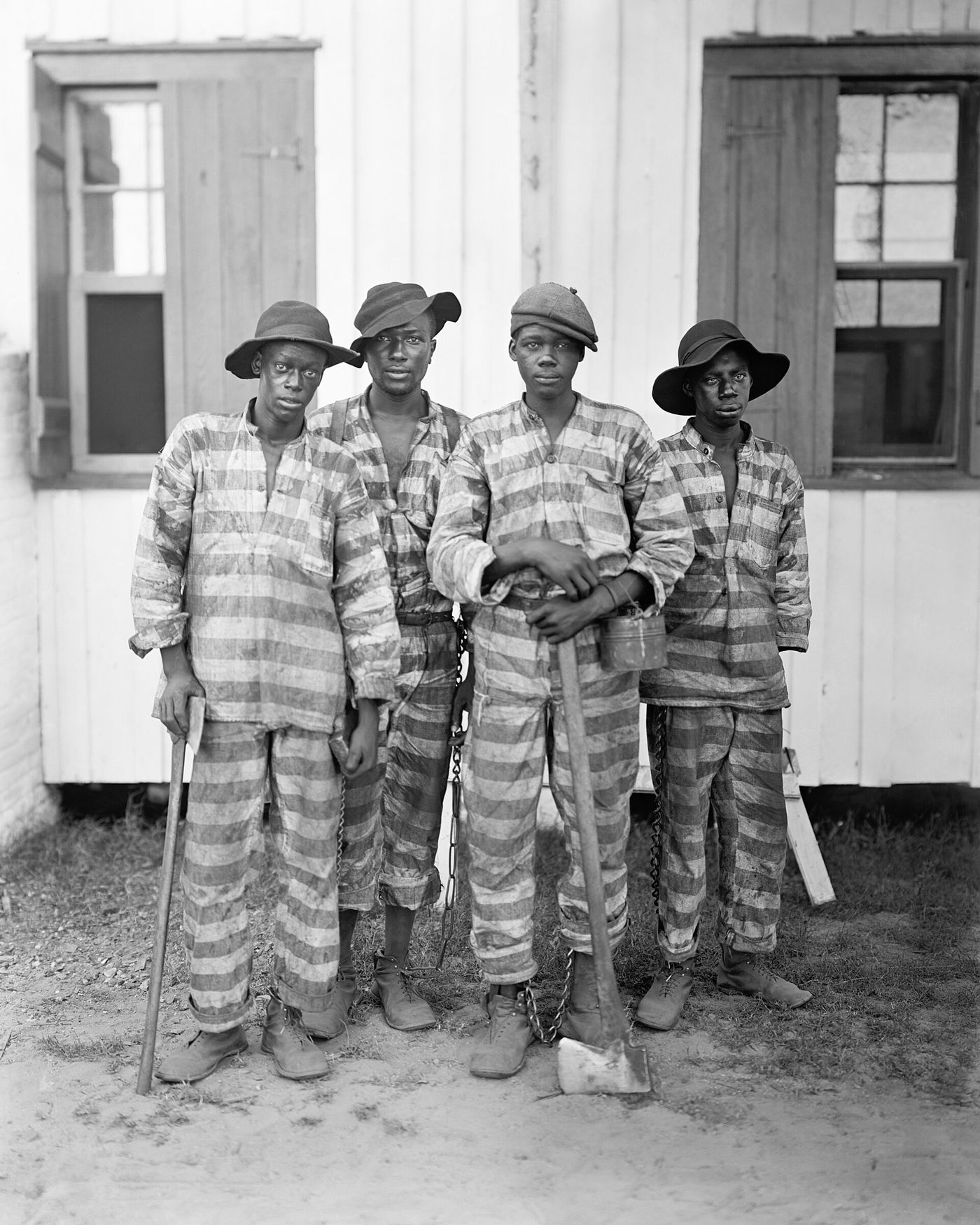 Four young men in striped prison uniforms standing with shovels beside a wooden building, about 1903