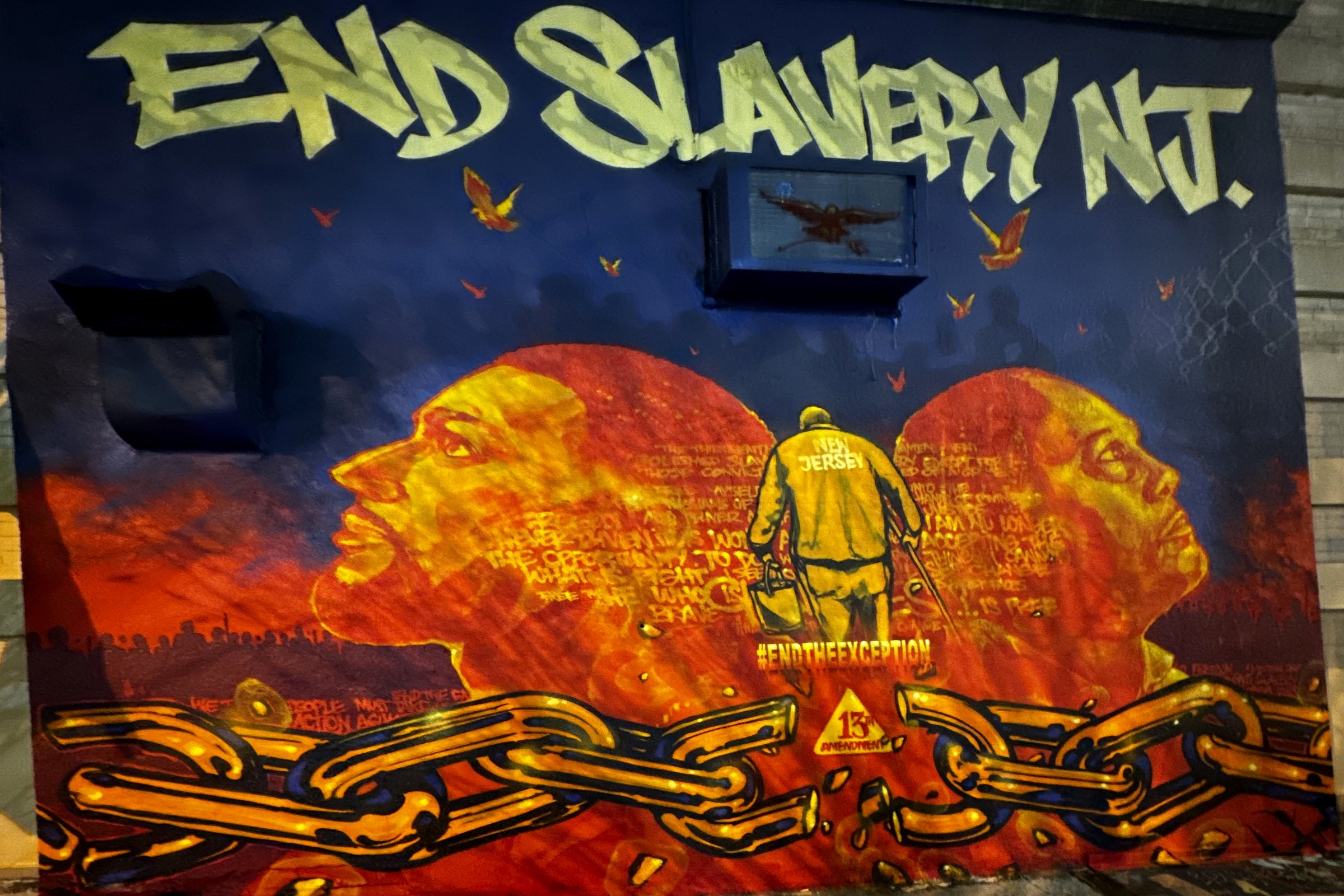 The End Slavery NJ mural: two profiles in red and gold, a figure in a New Jersey prison uniform walking away, and a broken chain across the bottom