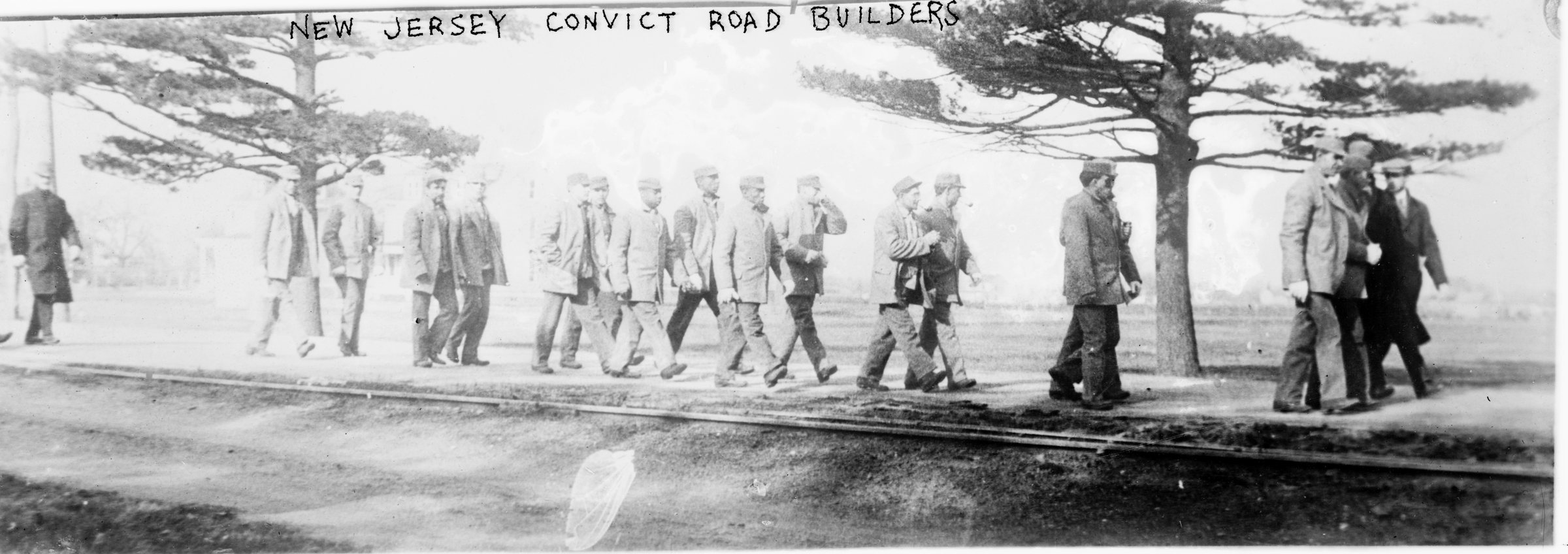 Una fila de convictos con ropa de trabajo marcha por un camino bajo árboles desnudos; el negativo lleva la leyenda New Jersey Convict Road Builders
